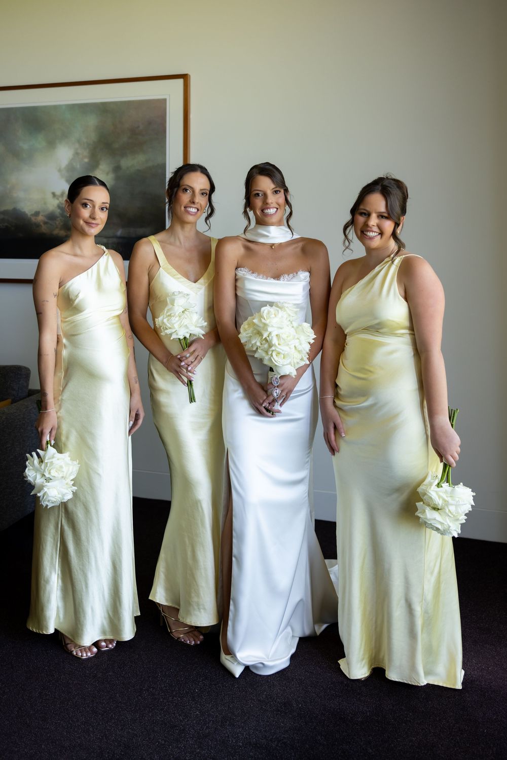 Bridesmaids pose together in pale yellow and white dresses holding white bouquets at a wedding.