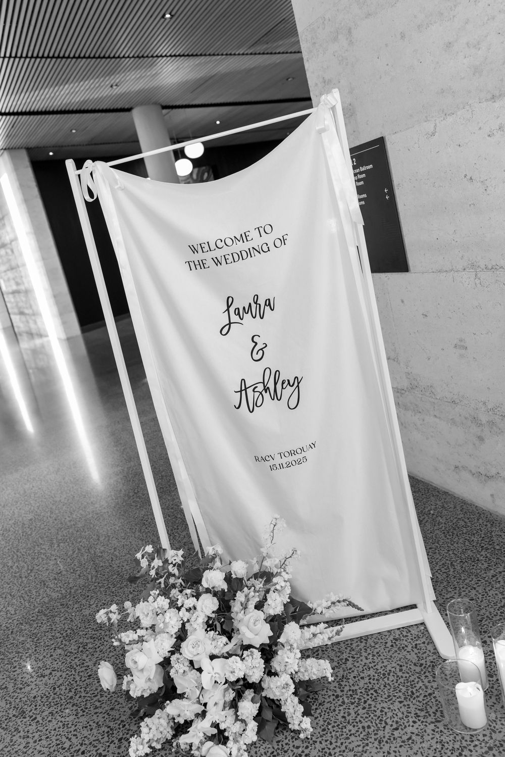 A white wedding welcome banner with floral decorations and candles displayed at the entrance of a venue.
