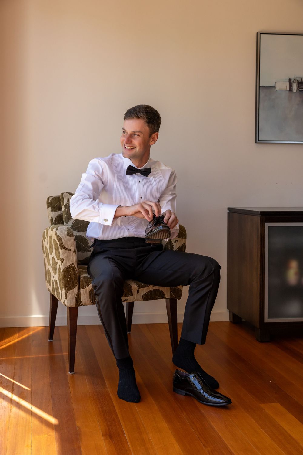 Formally dressed person in white shirt and bow tie sits relaxed in an upholstered armchair on hardwood floor.