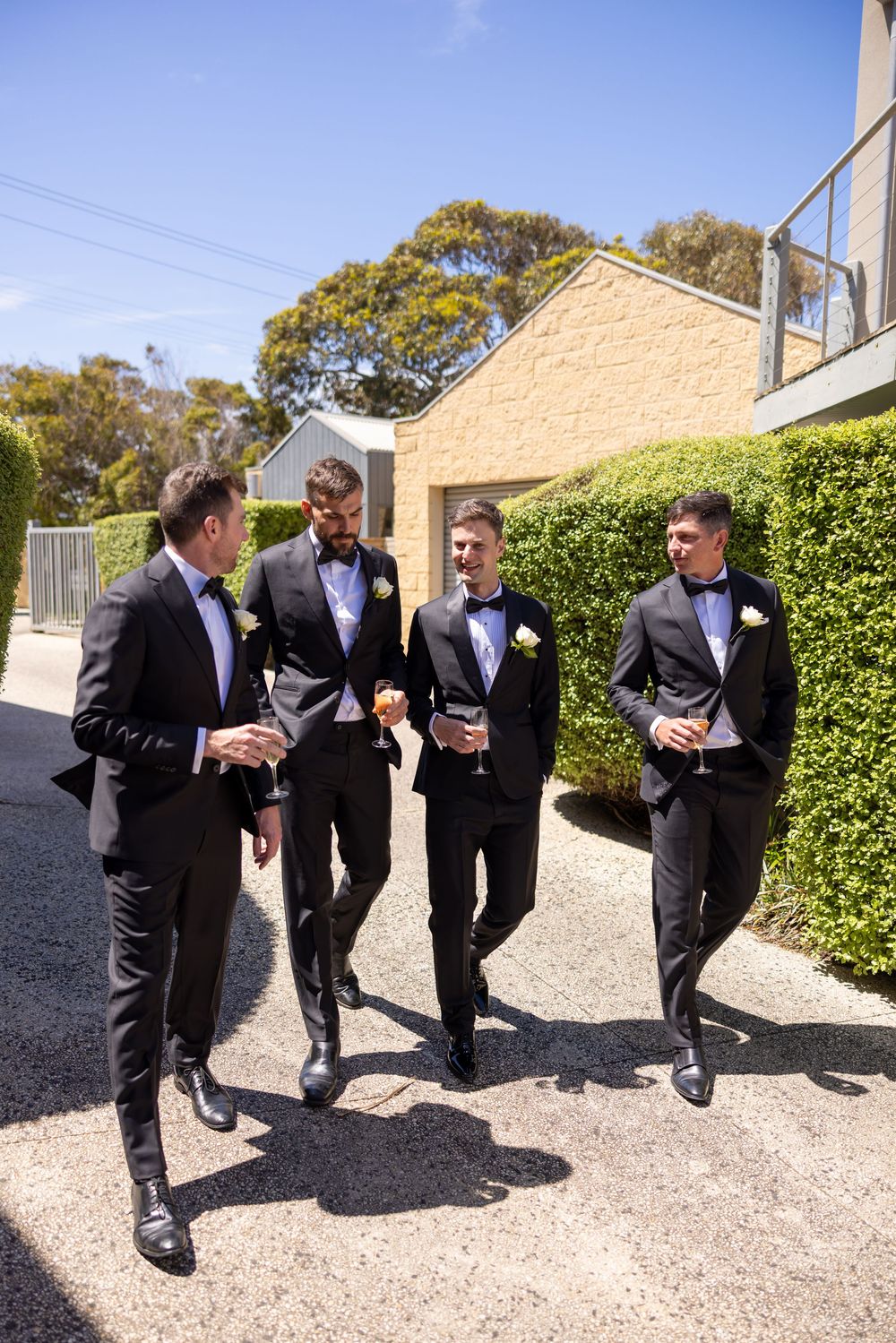 Groomsmen in black tuxedos share a candid moment outdoors during a wedding celebration.