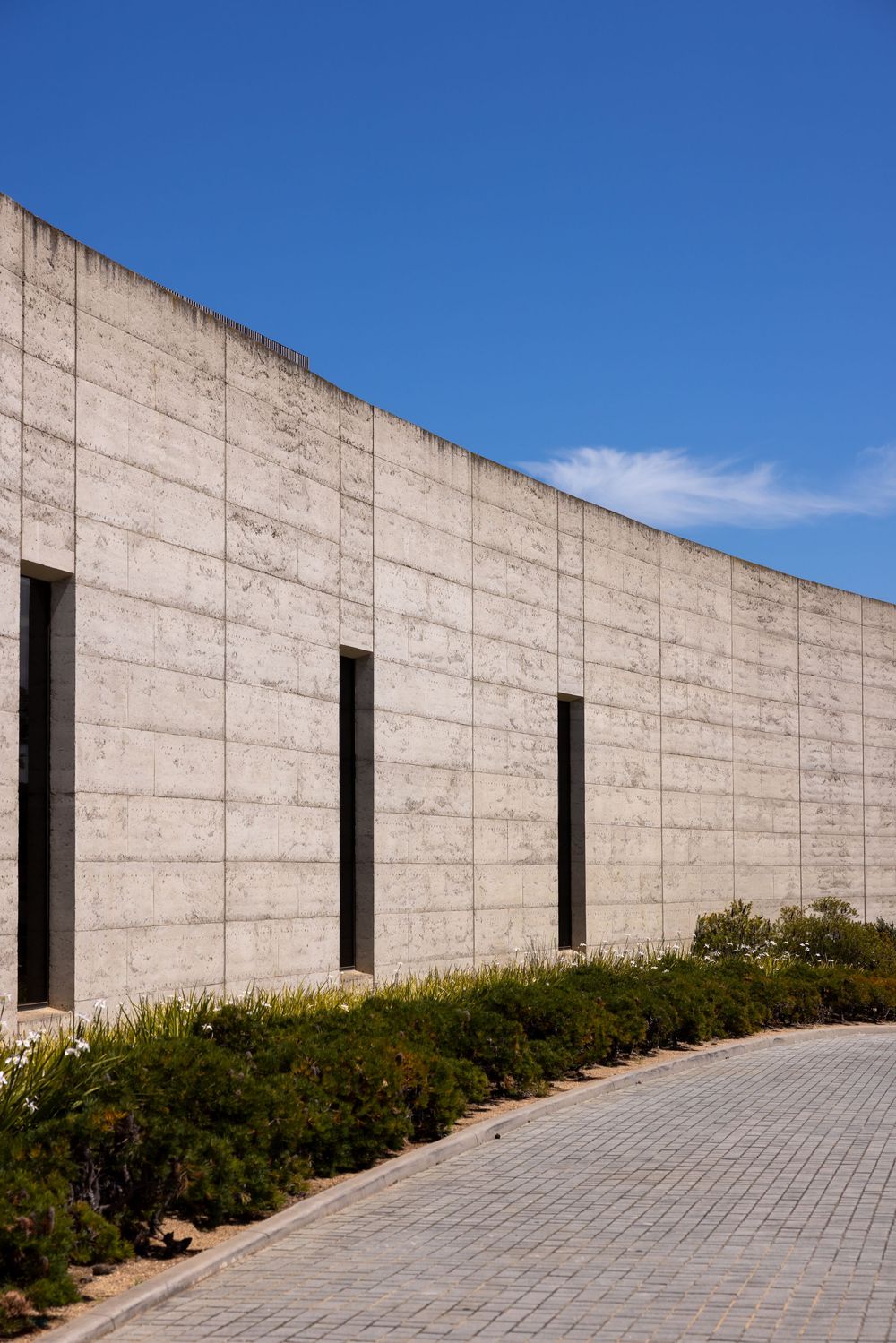 Modern concrete wall with vertical windows against blue sky and landscaped path.