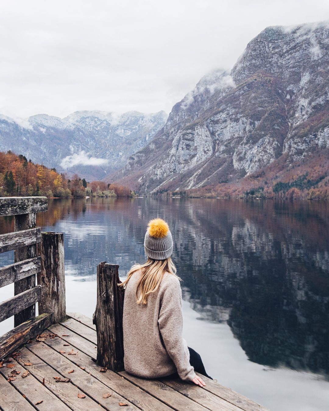 Person in winter attire sits on wooden dock overlooking misty mountain lake with autumn colors and snow-capped peaks.