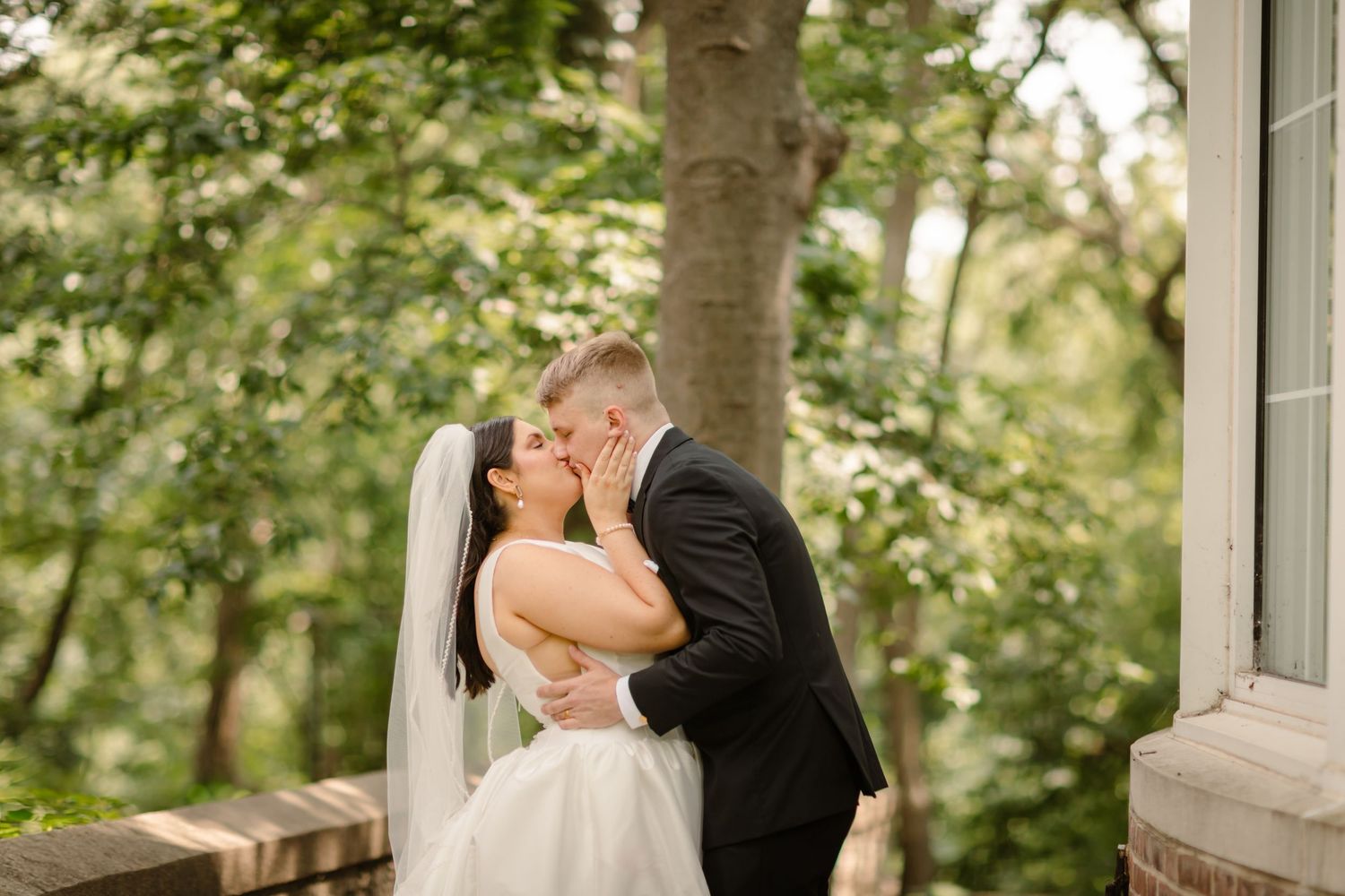 A romantic wedding couple shares a kiss outdoors surrounded by lush green trees and natural scenery.