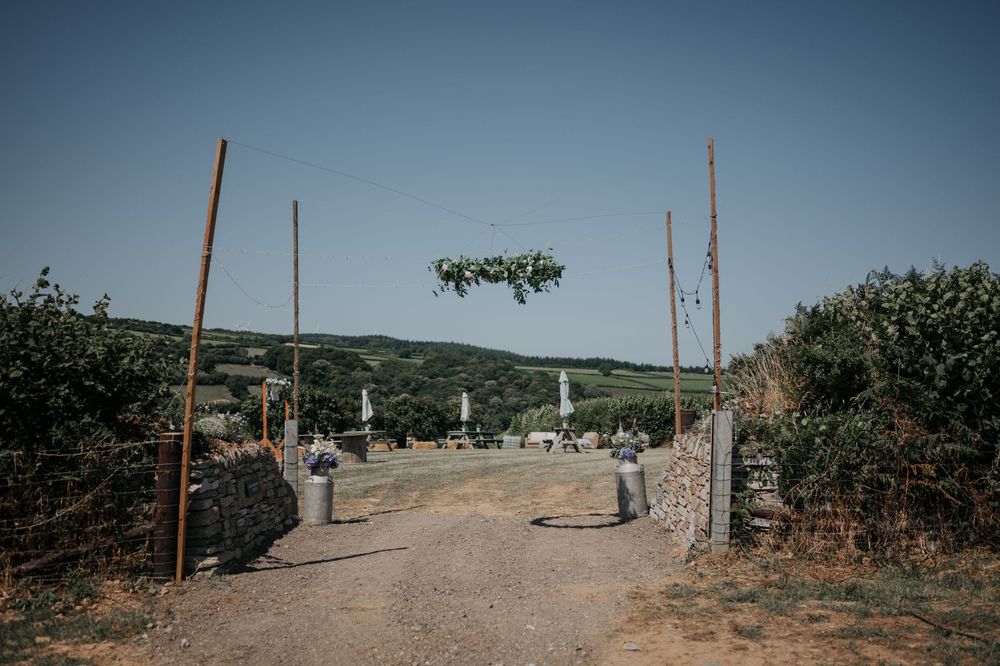 Rustic outdoor wedding venue entrance with hanging floral arch and stone walls under a clear blue sky.