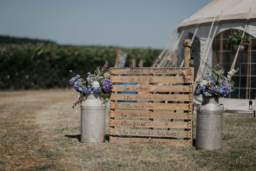 Rustic wedding welcome sign on wooden crate flanked by vintage milk churns with blue wildflowers.