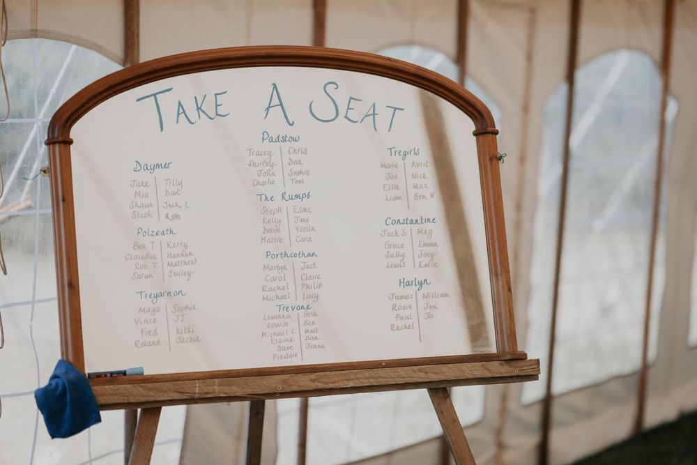 Elegant wedding seating chart on a wooden easel displaying table assignments in a cream marquee tent.