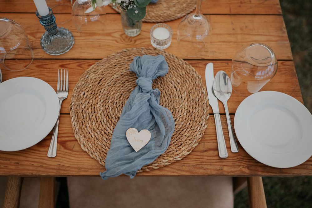 Rustic wedding table setting with wicker placemats, blue linen napkins, and heart-shaped wooden name tags.