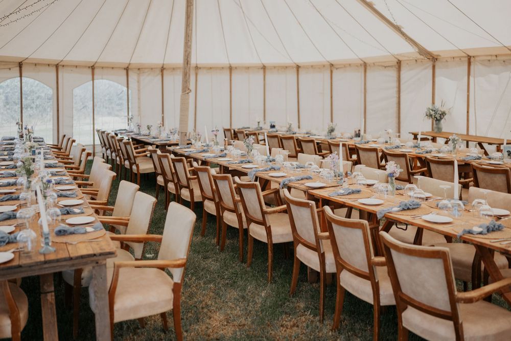Elegant marquee wedding reception with long wooden tables, upholstered chairs, and fairy light decor.