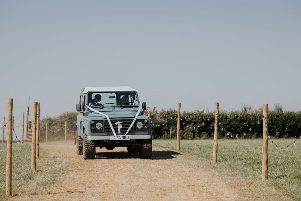 Vintage blue Land Rover Defender decorated with a white bow driving along a rustic countryside path.