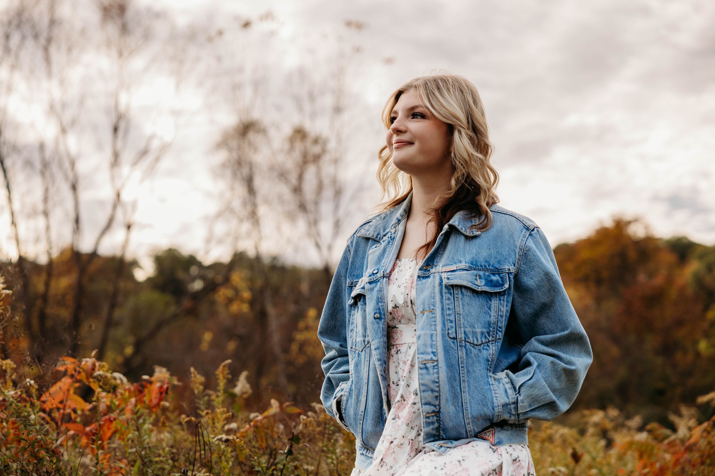 Woman in denim jacket and floral dress poses in field with autumn trees and cloudy sky in background.