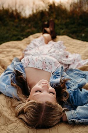 A woman in a floral dress and denim jacket lies peacefully on a blanket outdoors during golden hour.