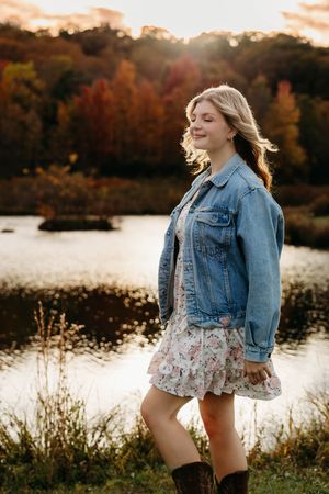 A person in a denim jacket and floral dress poses by a lake during golden hour with autumn foliage in the background.