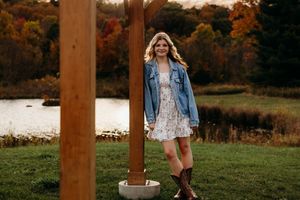 A person in a floral dress and denim jacket poses by a wooden post near a lake during autumn.