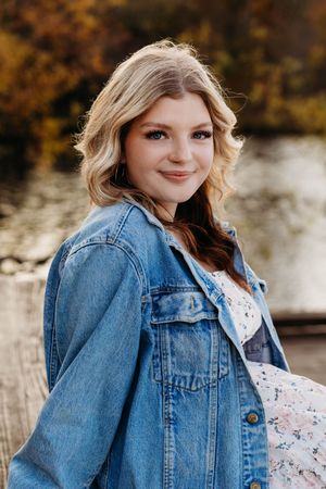 A person wearing a denim jacket and floral dress poses outdoors during autumn with a blurred natural background.