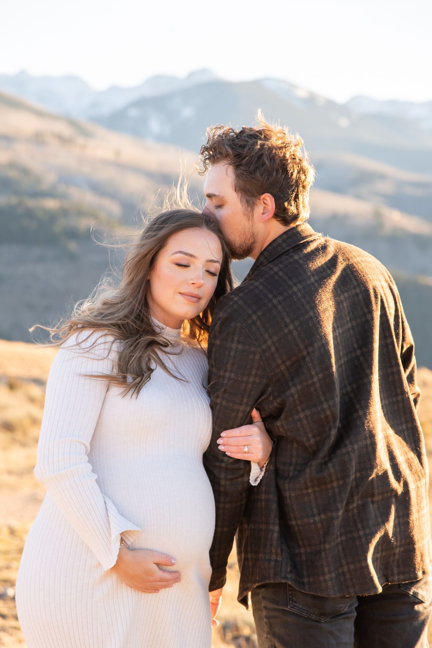A stylish couple in white sweaters embraces during a sunset maternity photoshoot in Vail Colorado.