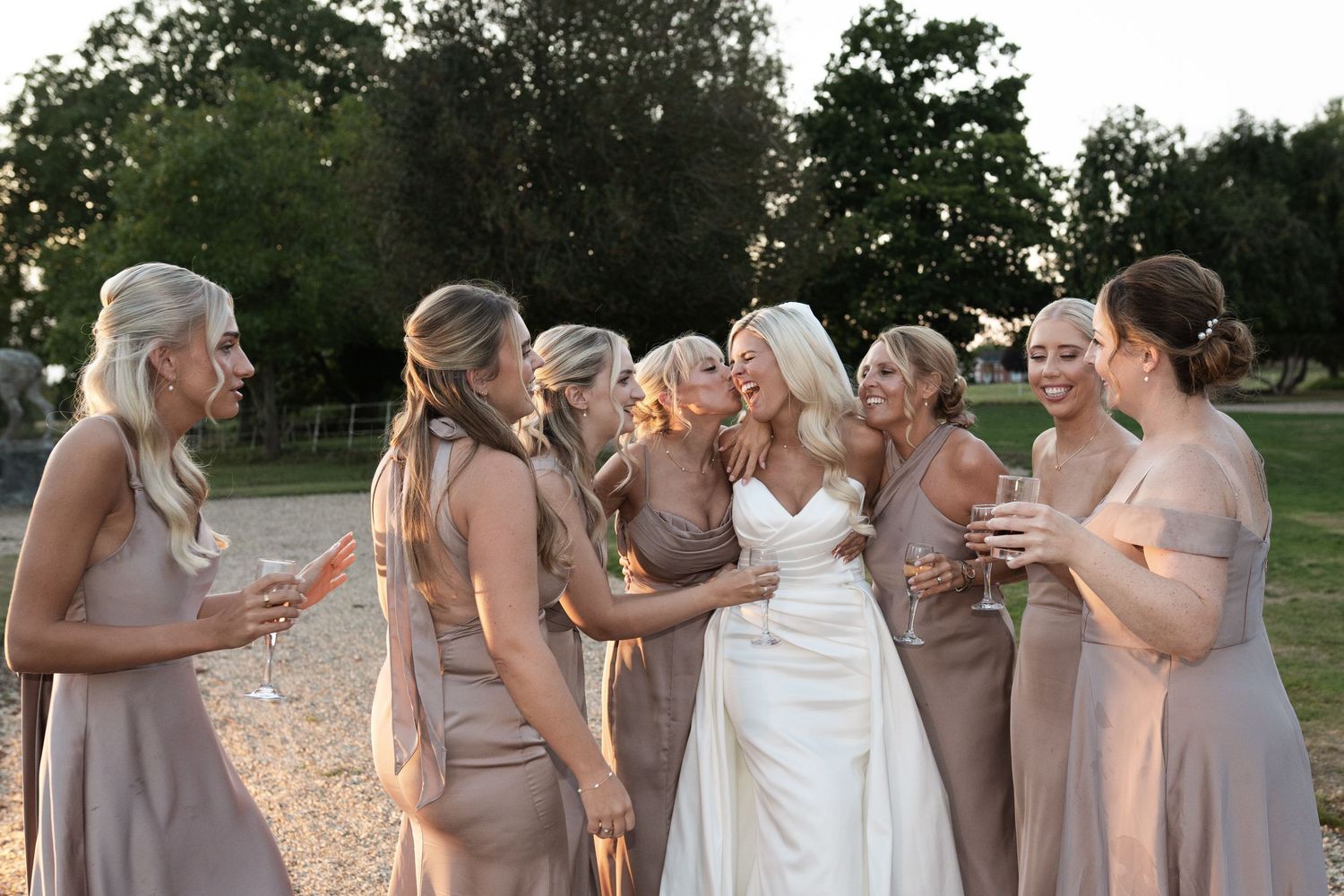 Wedding party in neutral-toned dresses sharing champagne toasts outdoors during golden hour.