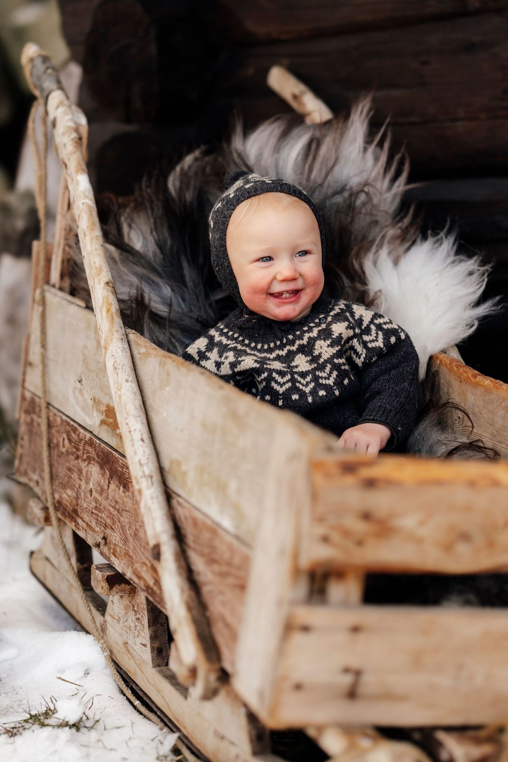 A smiling baby wearing a Nordic sweater sits in a rustic wooden sleigh with white fur accents.
