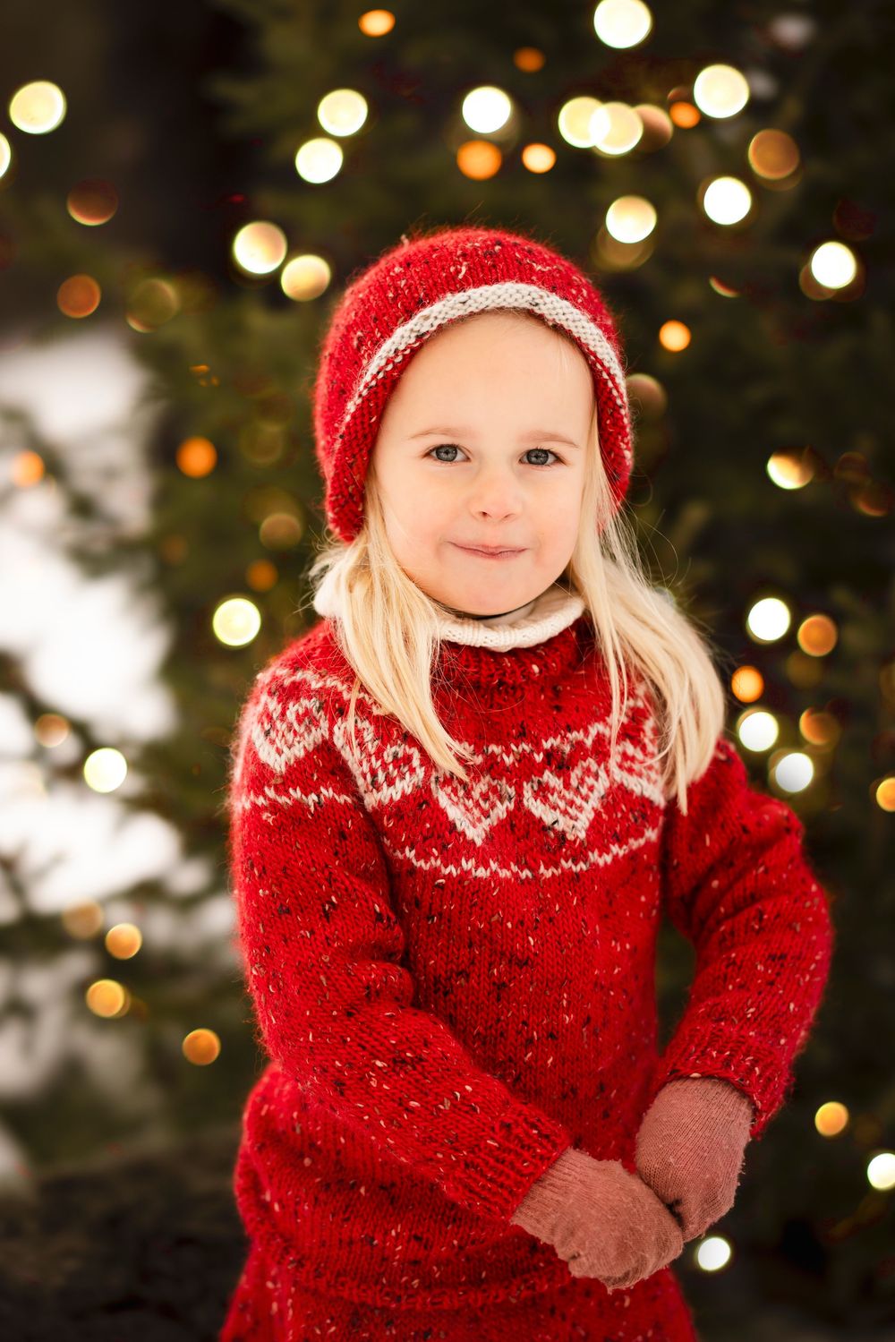 A child in a festive red Nordic sweater and matching hat stands in front of twinkling Christmas lights and evergreen branches.