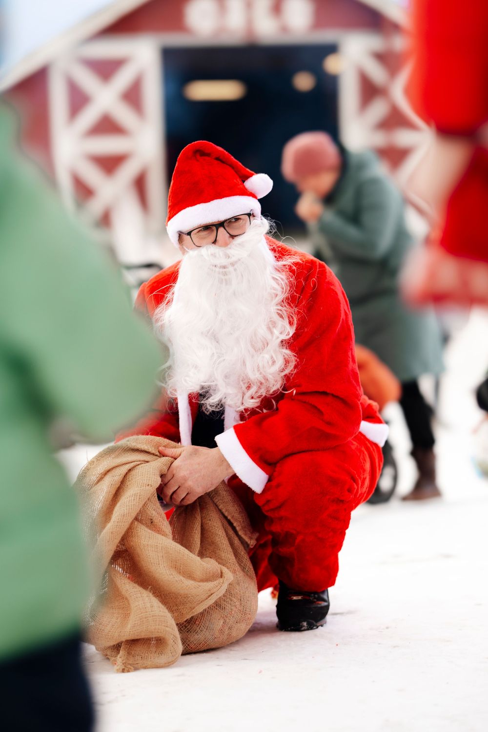 Santa Claus figure in traditional red suit sits with a large burlap gift sack in a holiday setting.