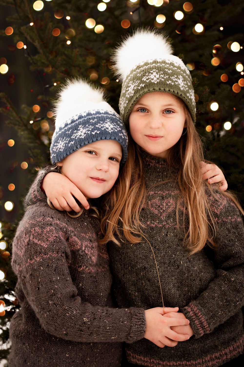 Two children wearing matching brown sweaters and winter hats pose together against a background of holiday lights.