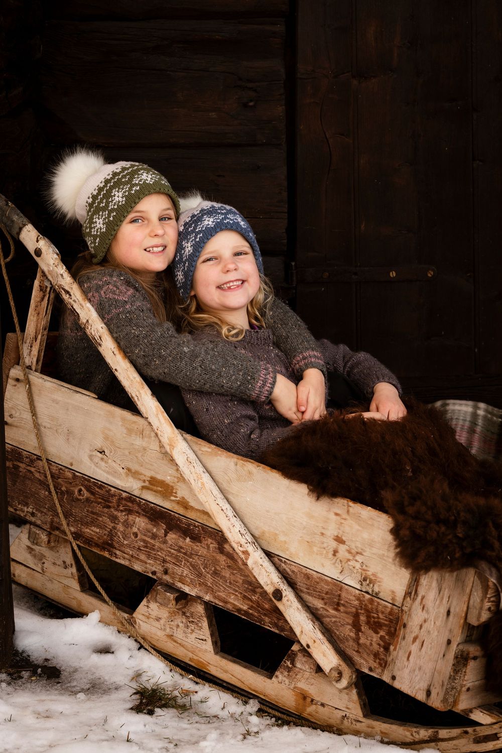 Two children in winter hats and sweaters sit together in a rustic wooden sleigh with fur blanket in snowy setting.