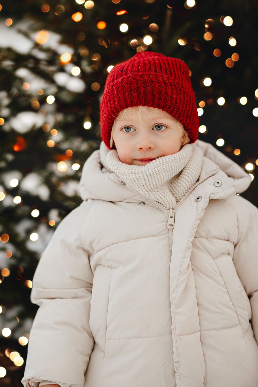 A child wearing a red knit beanie and cream puffer coat stands against a blurred background of holiday lights.