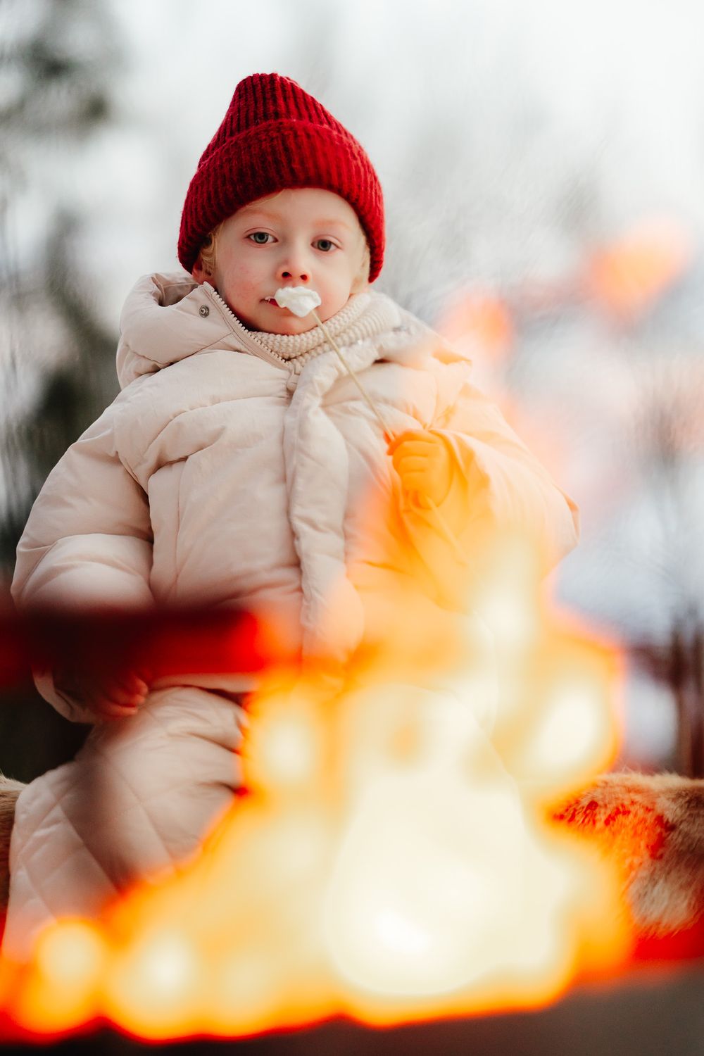 A small figure in a red beanie and cream puffer coat sits against a blurred orange fire in a winter setting.