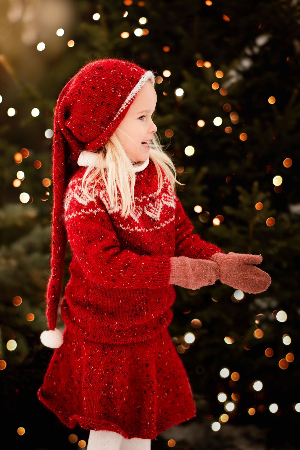 A child in a red knit sweater and hat plays joyfully against a backdrop of twinkling Christmas lights.