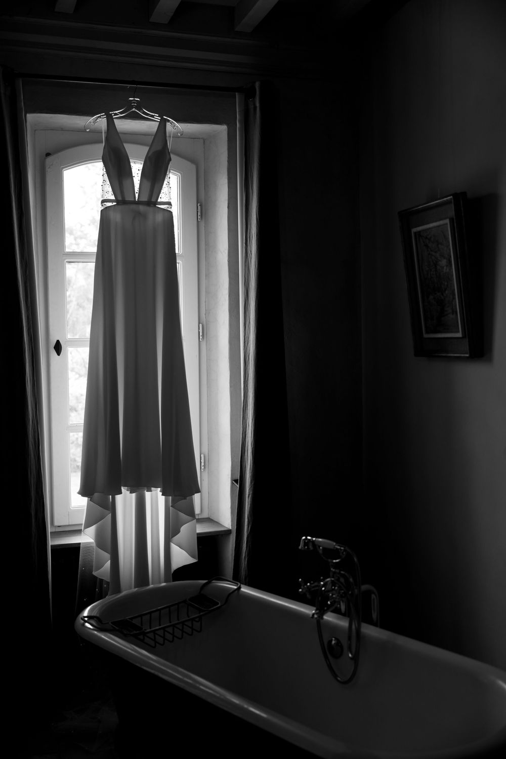 Wedding dress hangs in silhouette against bright windows in a dark room.