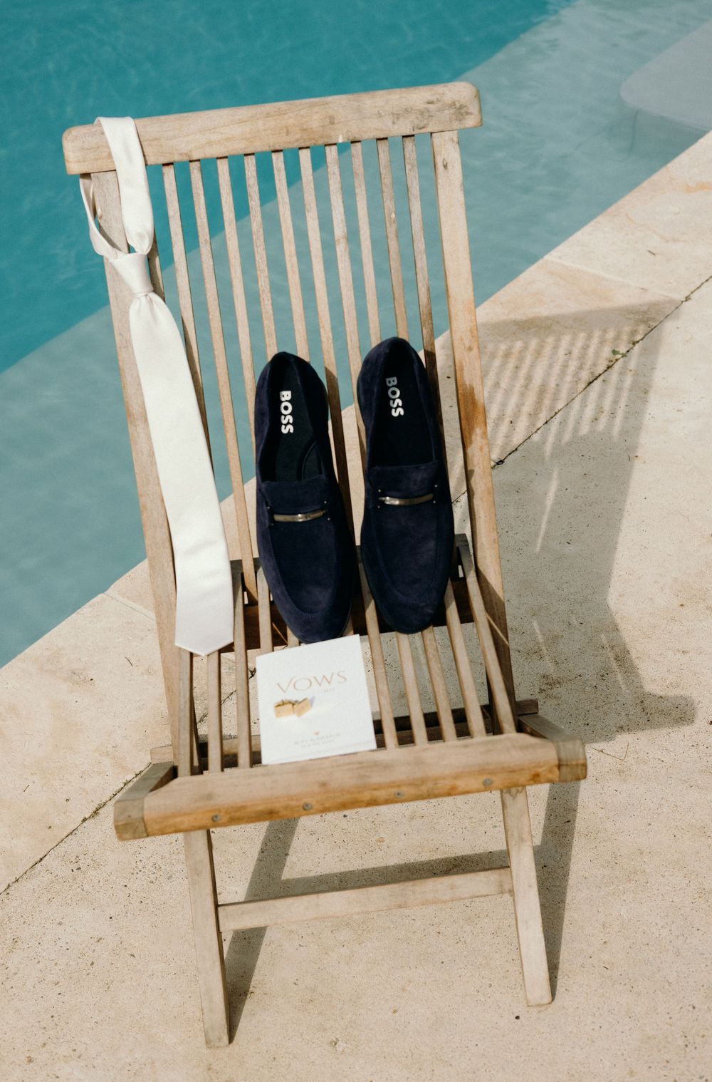 Wooden deck chair with formal shoes and clothing by a swimming pool.