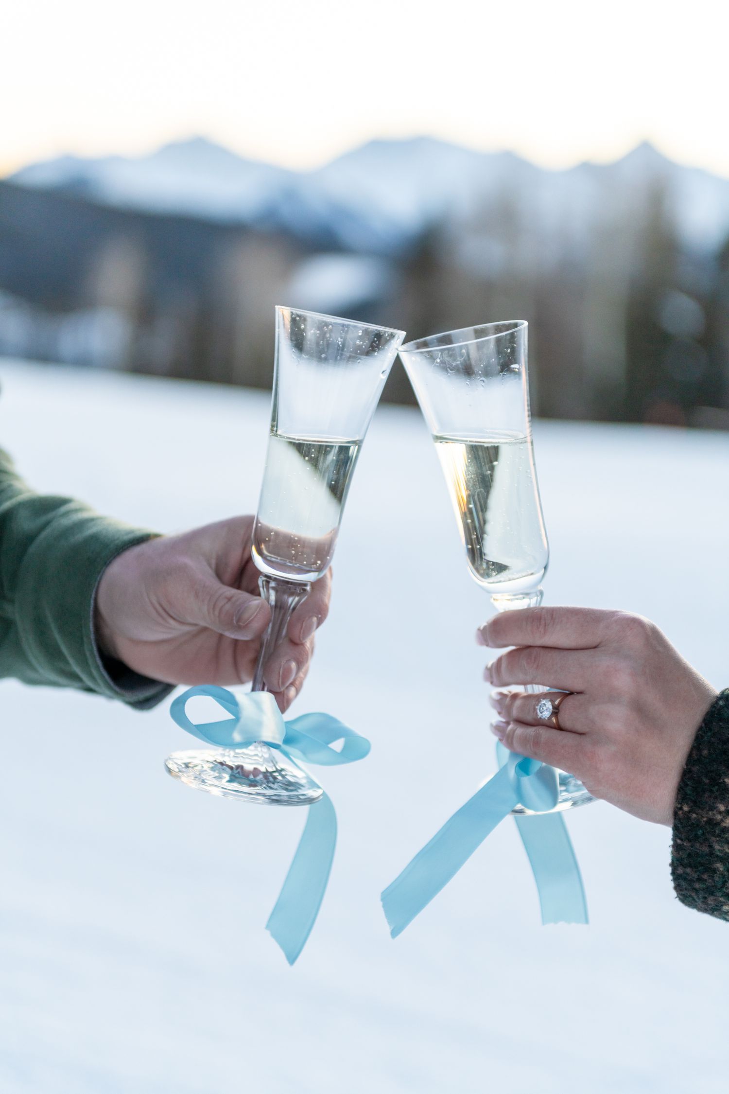 Two hands clinking champagne flutes with blue ribbons in a snowy mountain setting, one hand wearing an engagement ring.