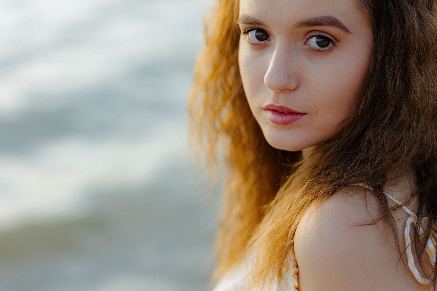 A woman with long red hair poses near the ocean at sunset, creating a dreamy golden-lit portrait.