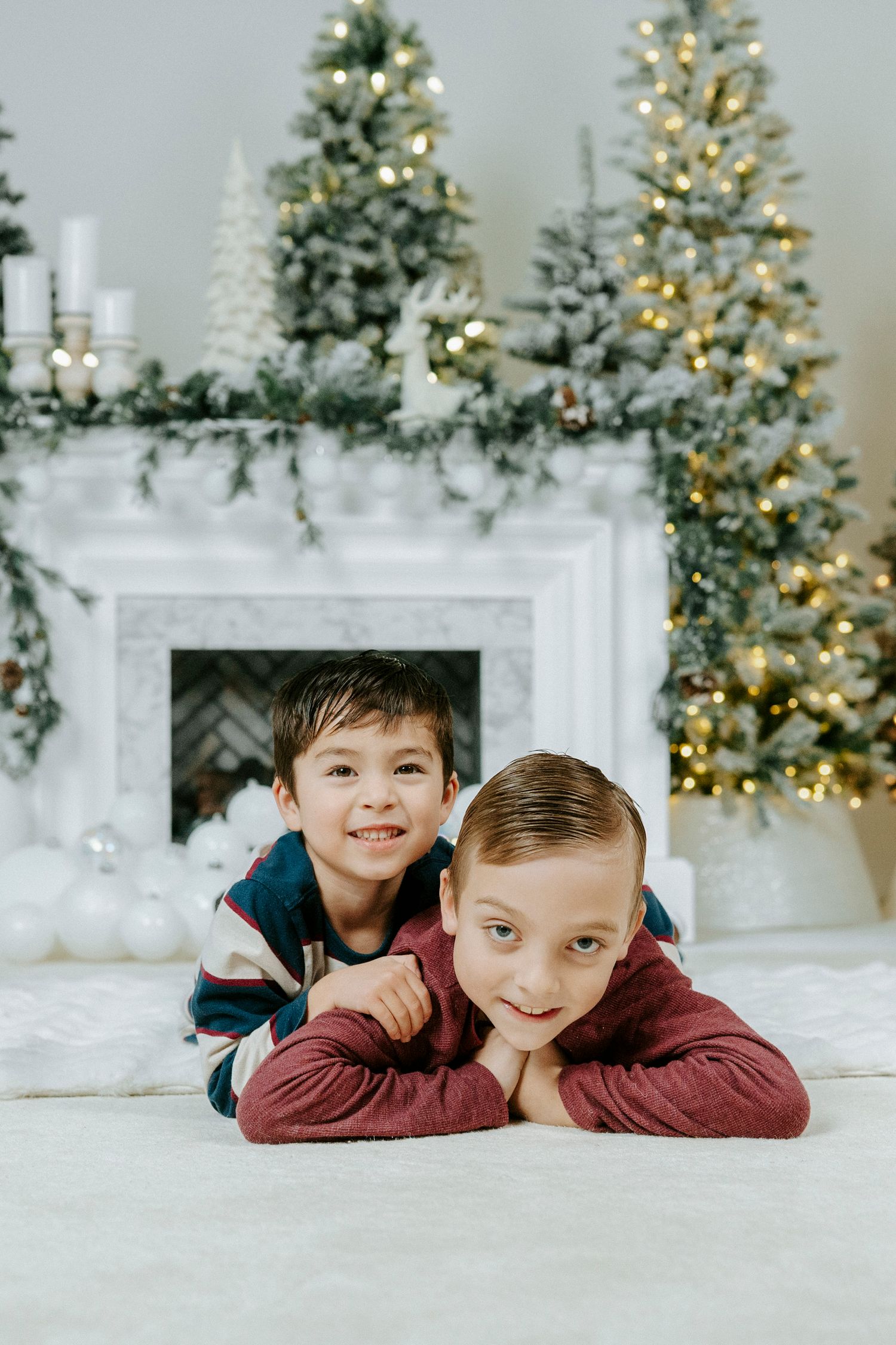 Two children in burgundy sweaters pose on white floor with decorated Christmas trees and mantel in background.