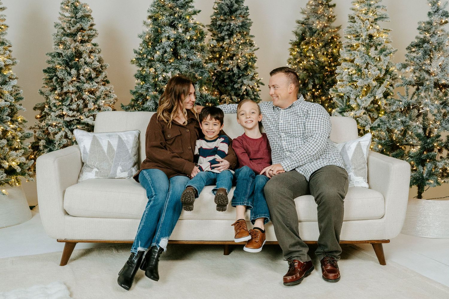 A family sits together on a white couch in front of multiple decorated Christmas trees in a bright studio setting.