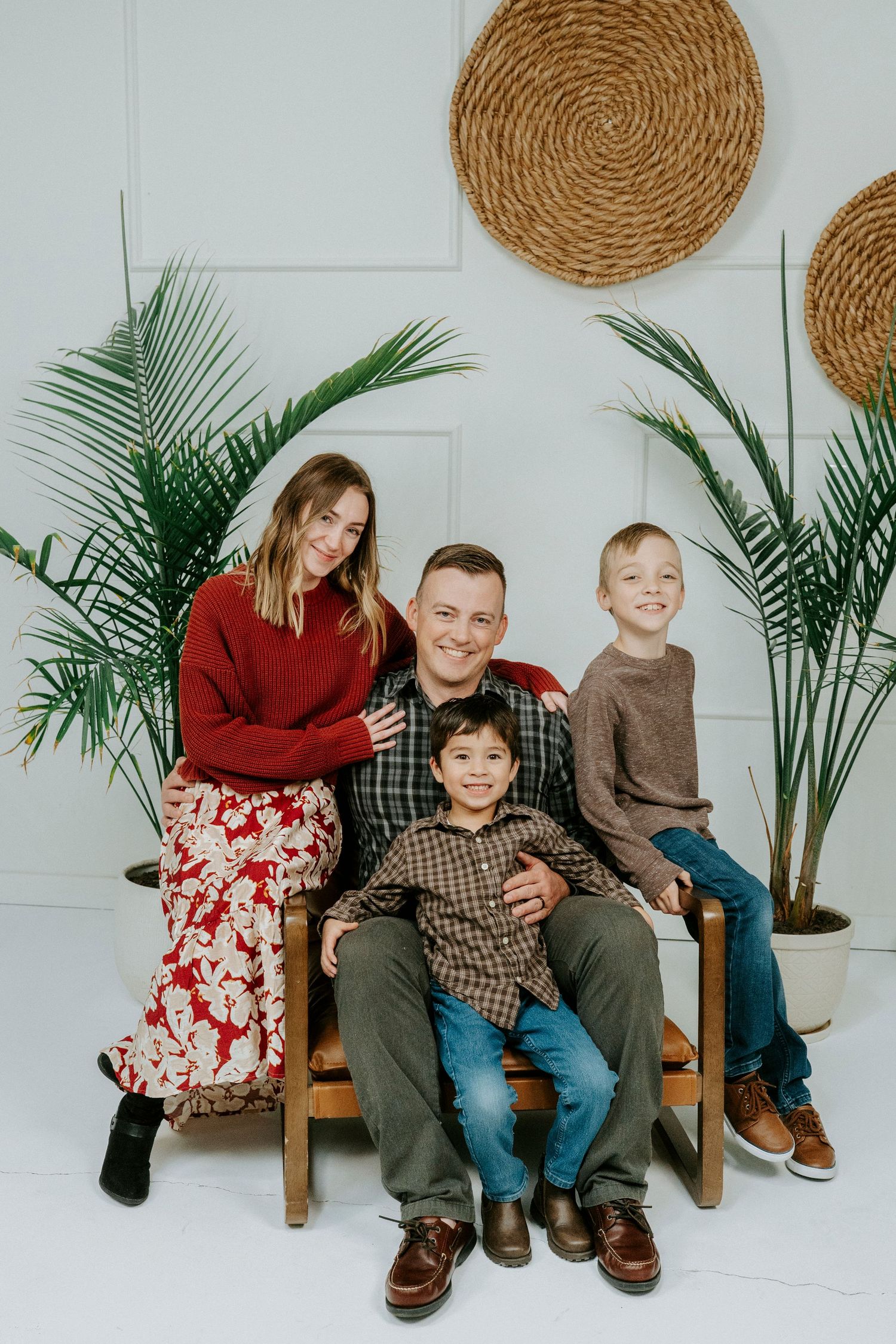 Family sits together on wooden bench with woven wall decor and tropical plants in minimalist white studio setting.