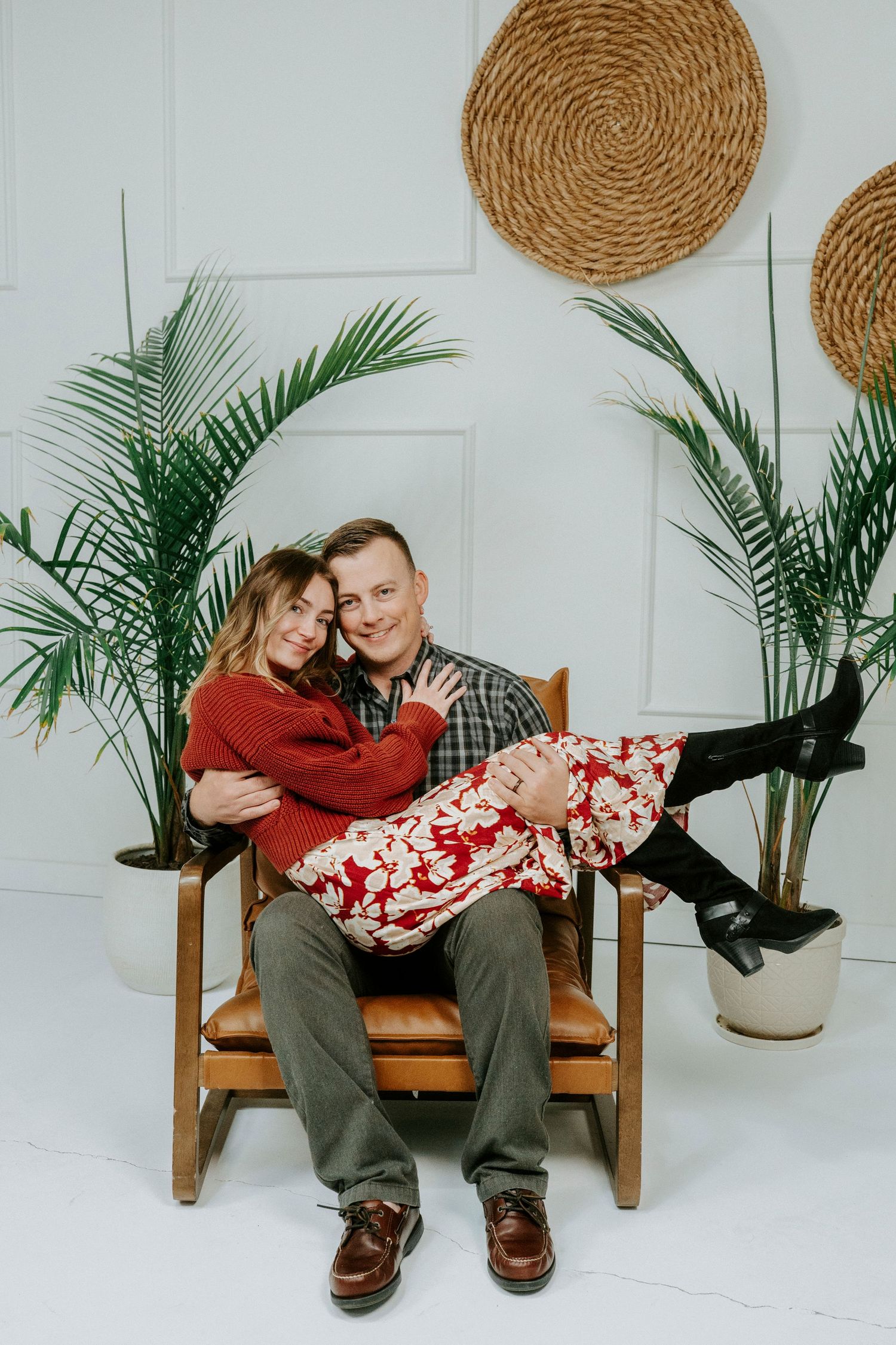 Couple sitting on wooden bench with woven wall decor and palm plants in minimalist white room.