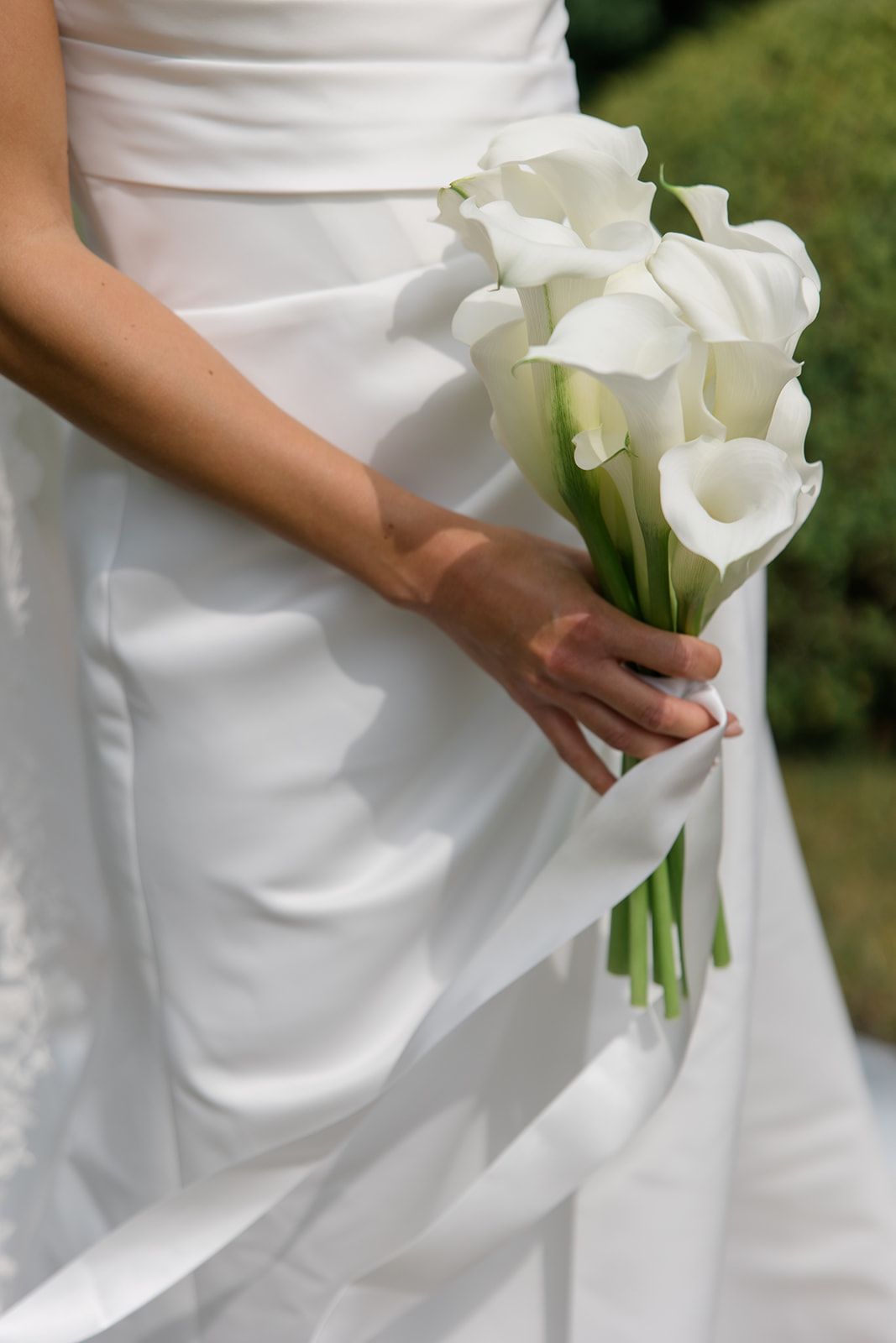 A white calla lily bridal bouquet held against an elegant white wedding dress in an outdoor garden setting.