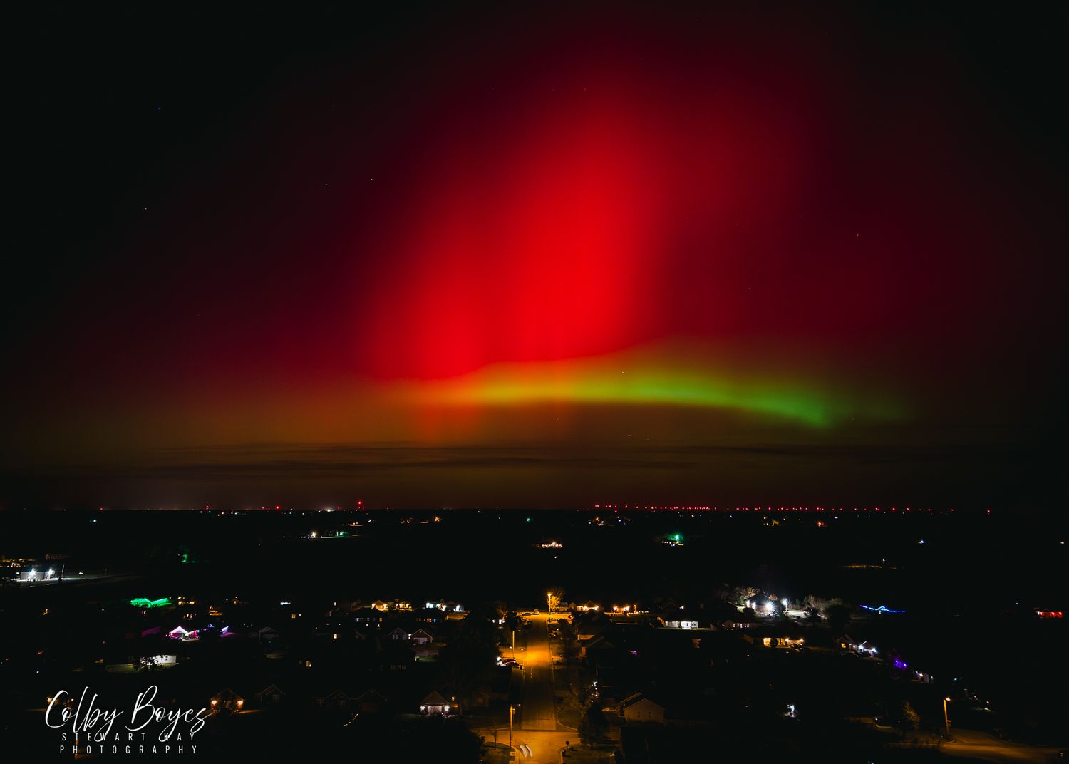 A stunning red and green aurora borealis lights up the night sky over a dark landscape with scattered lights below.