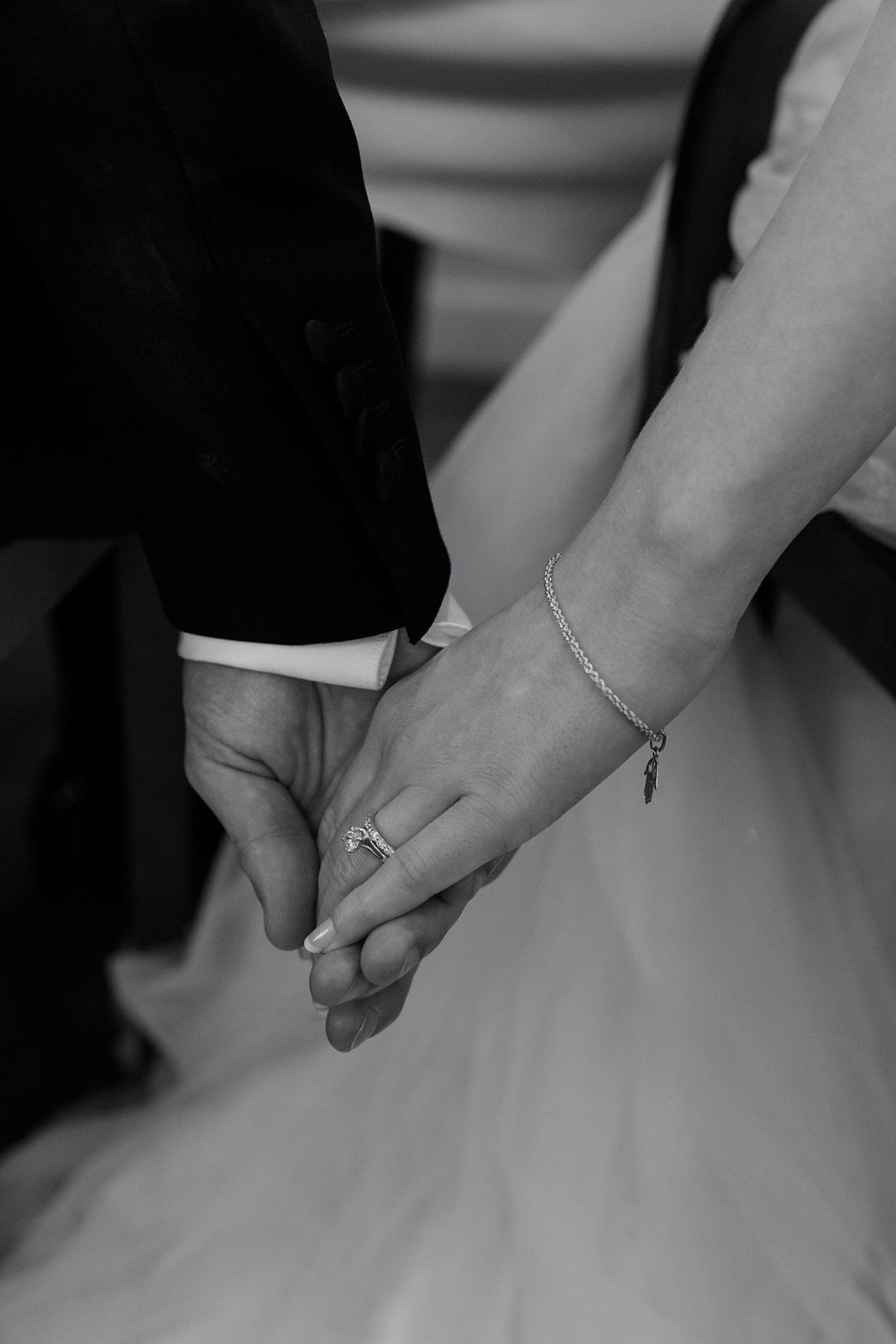 A black and white close-up of a couple holding hands, showing a wedding ring and delicate bracelet against a tulle dress.
