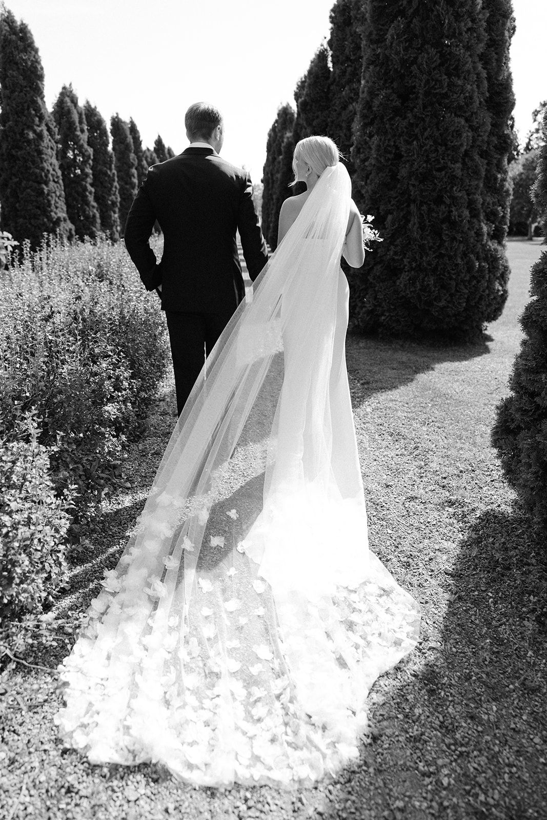 A black and white wedding photo showing a couple walking through a garden path lined with tall cypress trees.