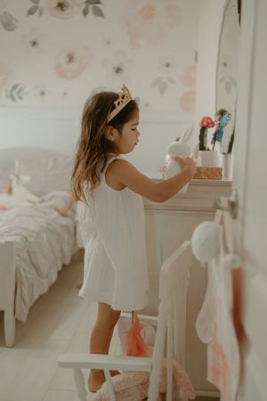 Child in white dress exploring bedroom with floral wallpaper and white furniture.