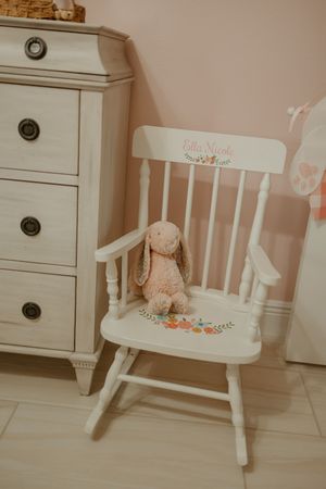 A white wooden rocking chair with a small teddy bear sits in a soft pink nursery room.