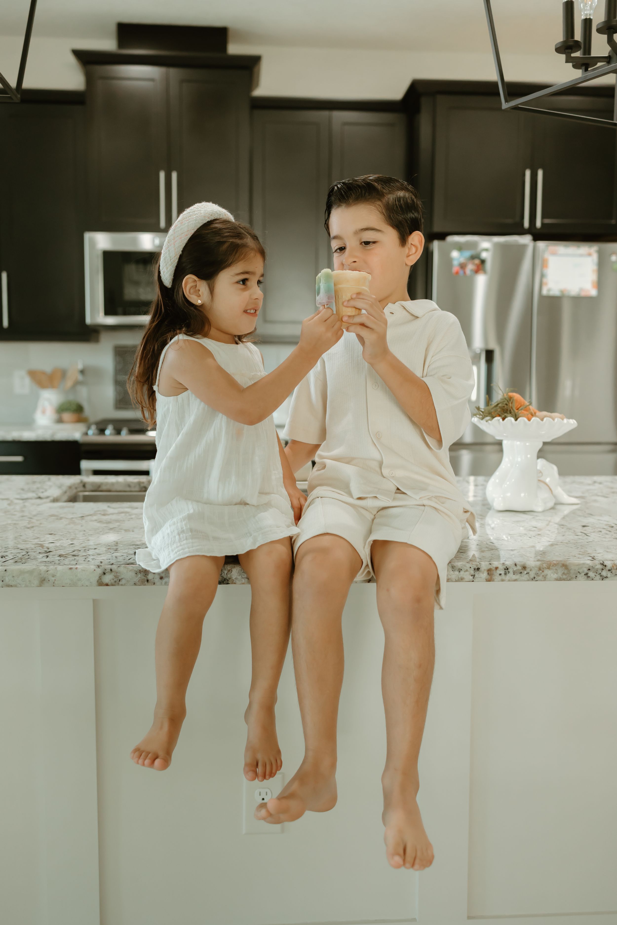 Children sitting on a kitchen counter with dangling feet in white outfits.