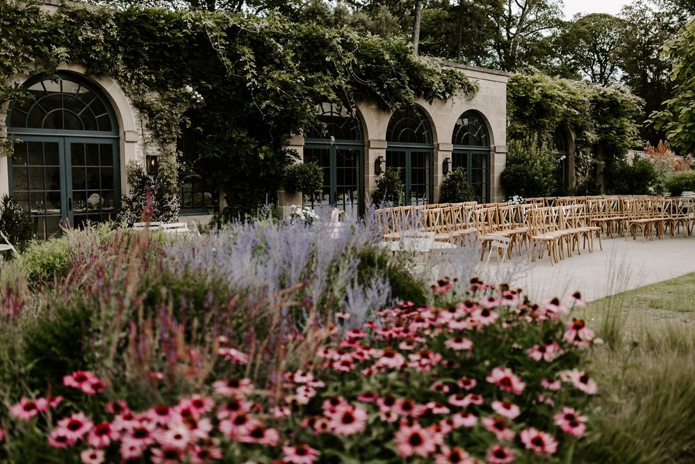 Beautiful garden venue with arched windows, pink flowers and lavender blooms alongside rows of wooden chairs for an event.