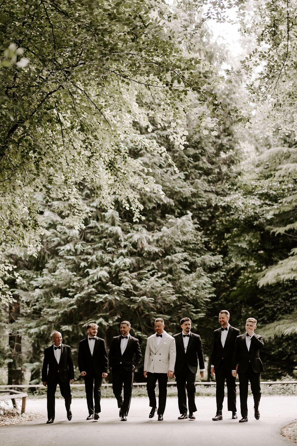 A wedding party in formal black tuxedos and one white jacket walks together on a tree-lined path in a natural setting.