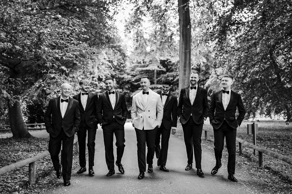 A wedding party in formal black and white attire poses together on a tree-lined path in a park setting.