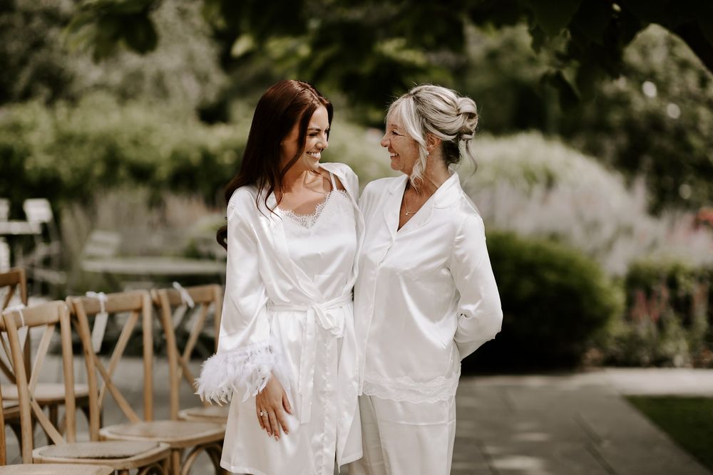 Two people in white outfits embrace and share a romantic moment outdoors on a rustic wooden bridge.