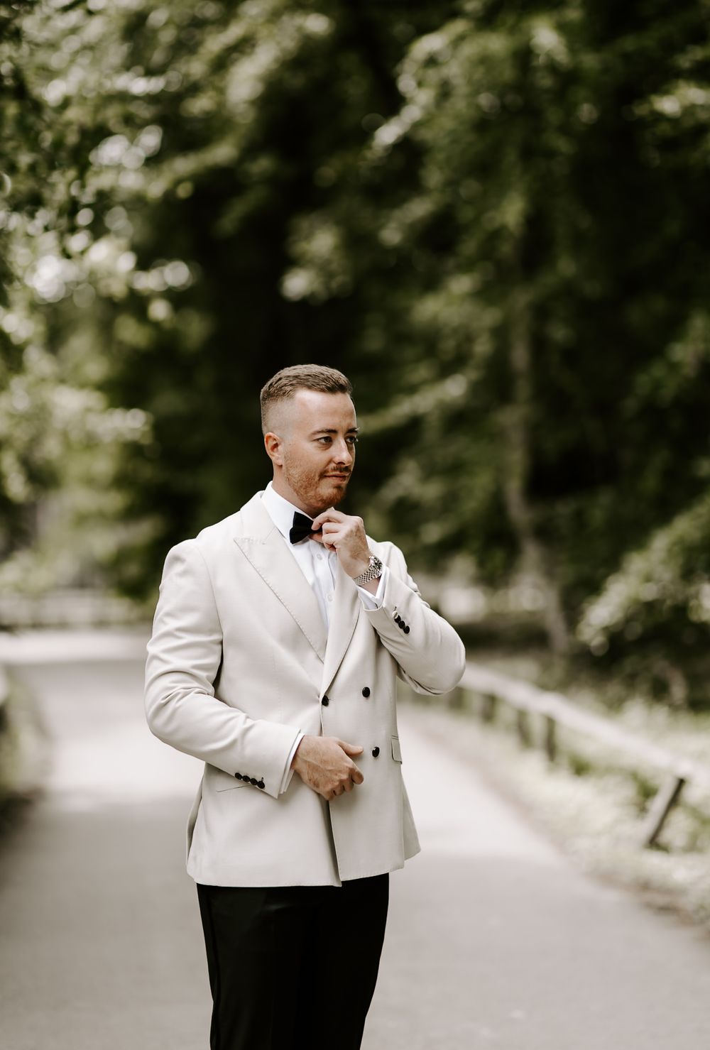 A person in a white suit jacket and black pants stands on a path surrounded by lush greenery.