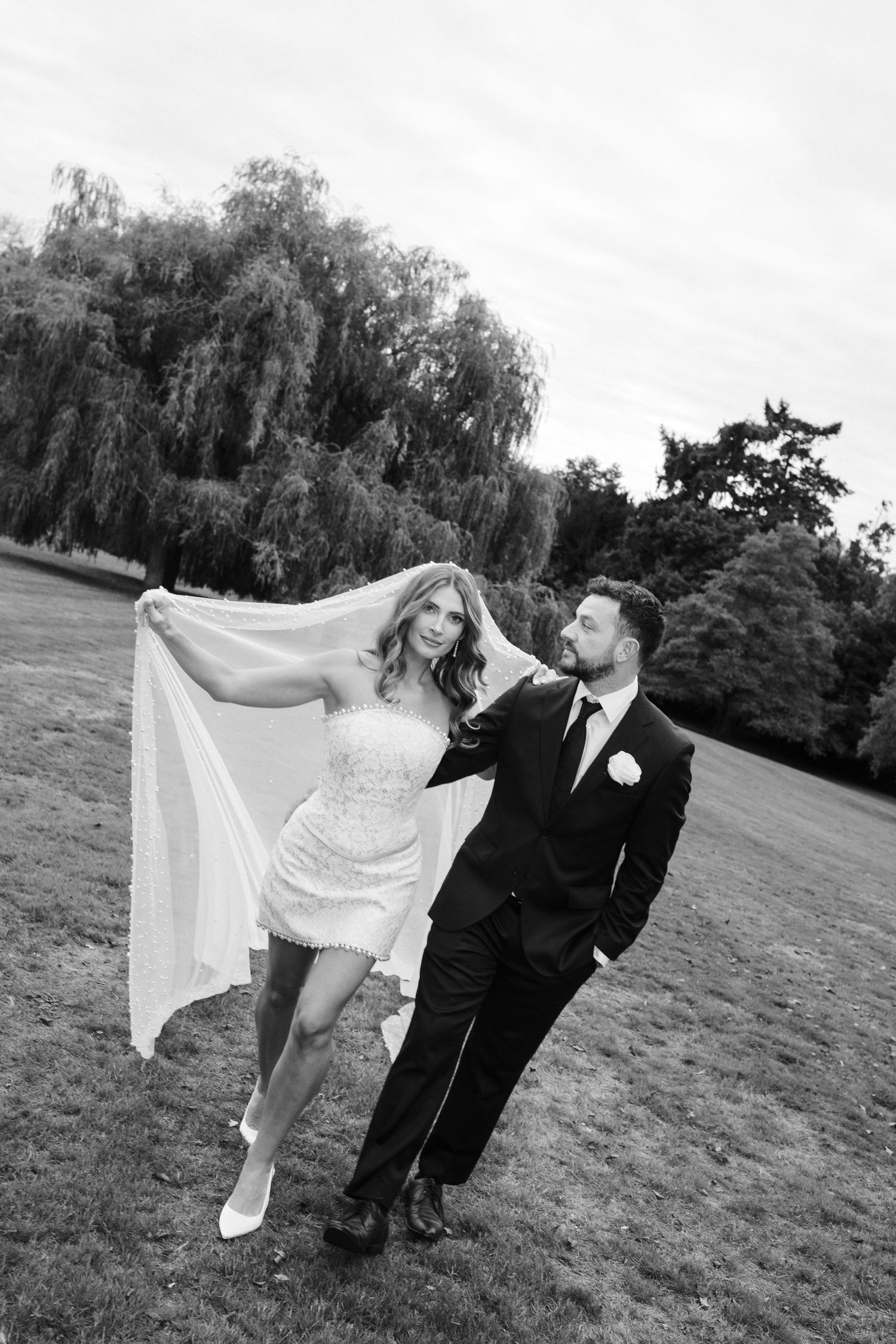 A couple in wedding attire playfully poses together on a grassy field with flowing veil movement in black and white.