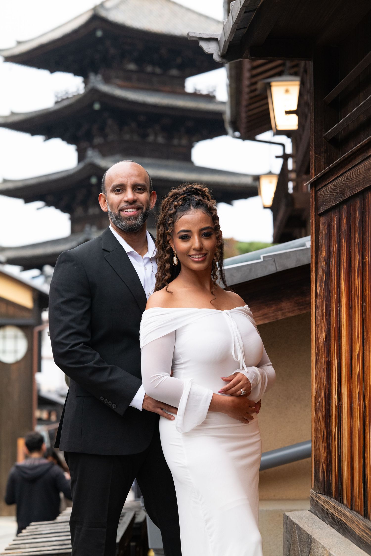 Couple poses by Yasaka Pagoda in Kyoto, woman in white off-shoulder dress, man in black suit.